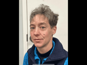 A person with short gray hair and a mole on their left cheek is wearing a blue shirt and navy vest, standing in front of a white door.