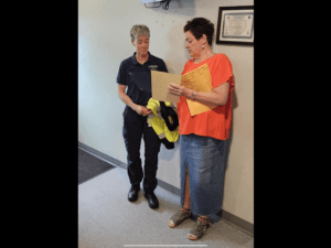 Two women stand indoors; one in a uniform holds a yellow jacket while the other in an orange shirt and denim skirt reads from a document. A framed certificate hangs on the wall.