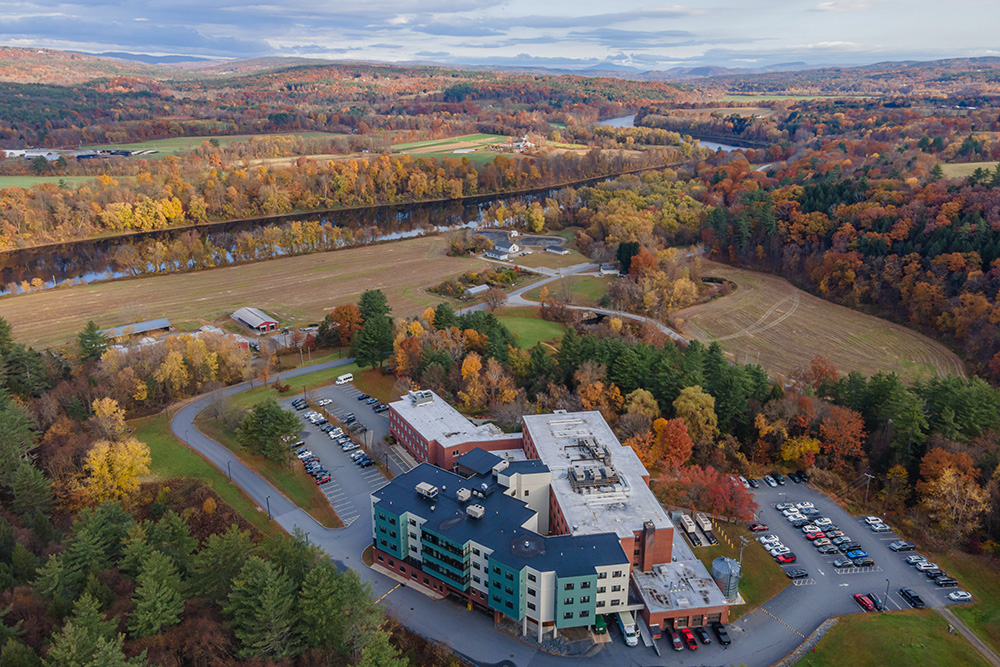 Aerial view of a large building complex with adjacent parking lots surrounded by autumn trees and fields, with a river and distant hills in the background.