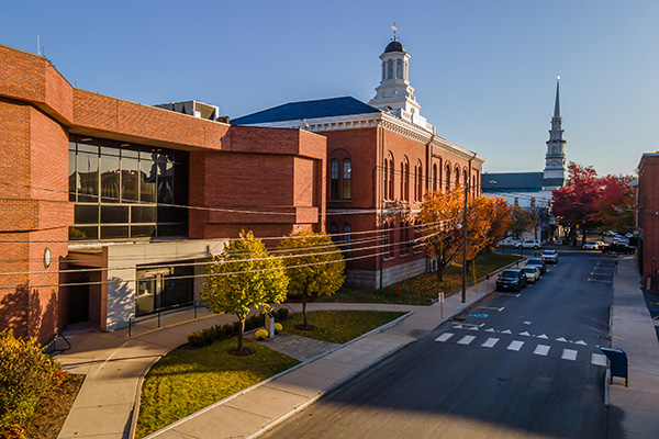 A street lined with brick buildings and autumn trees, with a prominent cupola and church steeple visible under a clear sky.