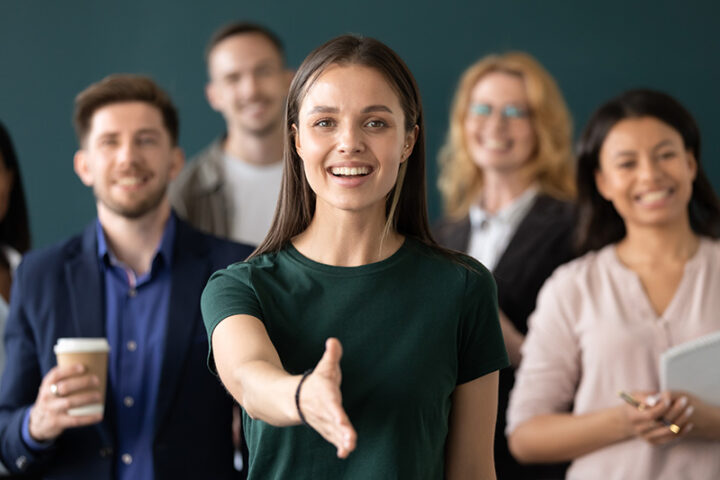 A woman in a green shirt extends her hand for a handshake, standing in front of a group of smiling people in business casual attire.