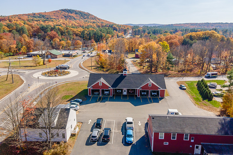 Aerial view of a fire station with four garage bays, surrounded by autumn trees, parked cars, and a nearby roundabout in a small town.