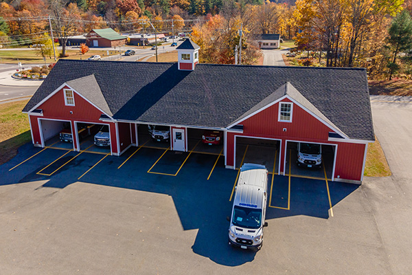 A red building with five garage bays, each containing a vehicle, and one white van parked in front; autumn trees and a road are in the background.