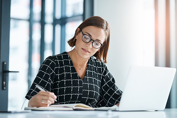 Woman with glasses working at a desk, writing in a notebook while using a laptop in a modern office setting.