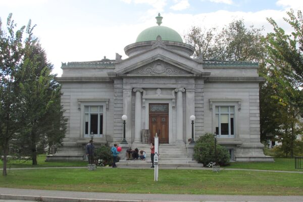 A small, neoclassical stone building with a green dome, columned entrance, and people gathered on the front steps, surrounded by lawns and trees.